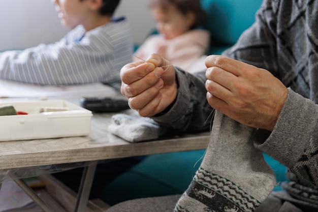 Mężczyzna szyjący skarpetę w salonie w domu, podczas gdy jego dzieci odrabiają lekcje