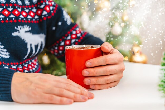 Mężczyzna trzyma kubek kakao z piankami przed choinką