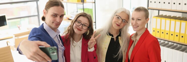 Zdjęcie mężczyzna trzyma smartfon i robi sobie selfie z koleżankami w biurze, zadowolonymi pracownikami w formie formalnej