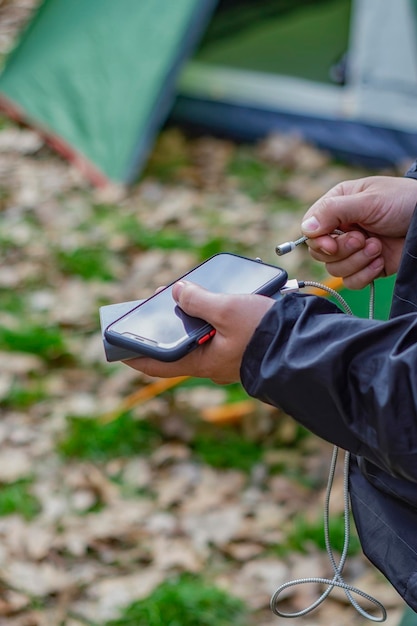 Mężczyzna trzyma w dłoniach smartfon i ładuje go powerbankiem na tle namiotu turystycznego w naturze Przenośna ładowarka podróżna