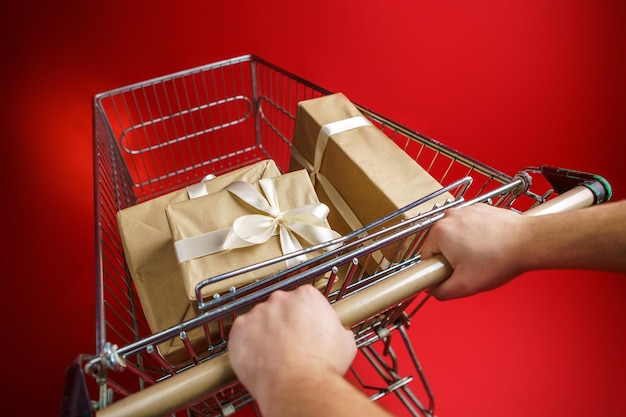 Mężczyzna trzyma w rękach wózek z supermarketu z prezentami owiniętymi w papier rzemieślniczy z białą wstążką na czerwonym tle