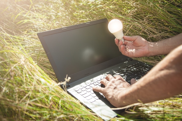 Mężczyzna trzyma żarówkę i używa laptopa w przyrodzie.