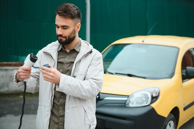 Mężczyzna trzymający kabel zasilający do samochodu elektrycznego na parkingu zewnętrznym I zamierza podłączyć samochód do stacji ładującej na parkingu w pobliżu centrum handlowego