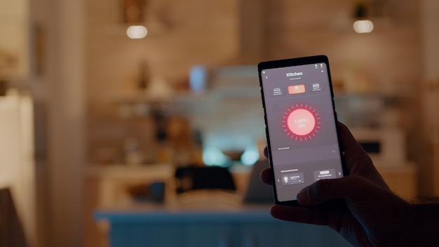 Mężczyzna trzymający smartfon z aplikacją do sterowania oświetleniem, włączający światła siedzący w kuchni z systemem automatyki oświetlenia. Osoba korzystająca z oprogramowania inteligentnego domu pracująca zdalnie przy laptopie