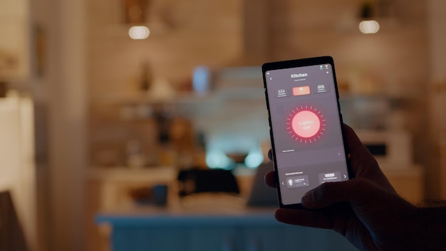 Mężczyzna trzymający smartfon z aplikacją do sterowania oświetleniem, włączający światła siedzący w kuchni z systemem automatyki oświetlenia. Osoba korzystająca z oprogramowania inteligentnego domu pracująca zdalnie przy laptopie