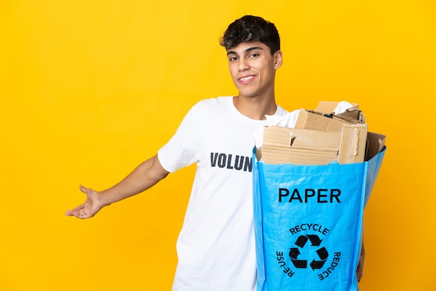 Mężczyzna trzymający worek do recyklingu pełen papieru do recyklingu na izolowanej żółtej ścianie wyciągając ręce na bok, by zaprosić do przyjścia