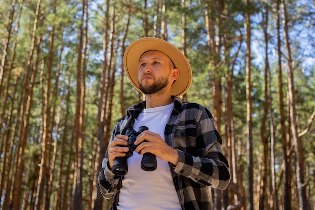 Mężczyzna Turysta W Kapeluszu I Koszuli W Kratę Spogląda Przez Lornetkę W Lesie