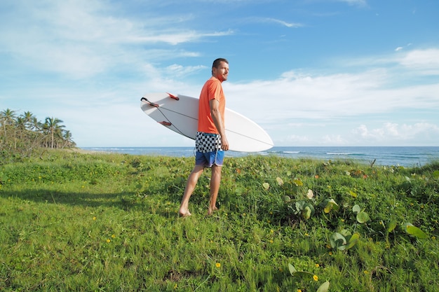 Zdjęcie mężczyzna ubrany pomarańczowy t-shirt i niebieski krótki gospodarstwa deska surfingowa chodzenie w trawie obok oceanu.