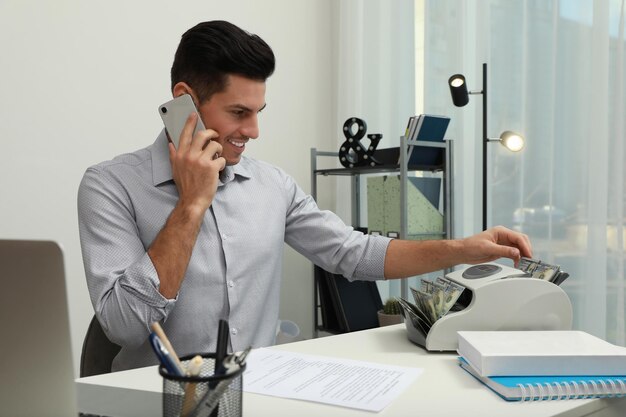 Mężczyzna używający licznika banknotów podczas rozmowy przez telefon przy białym stole w pomieszczeniu