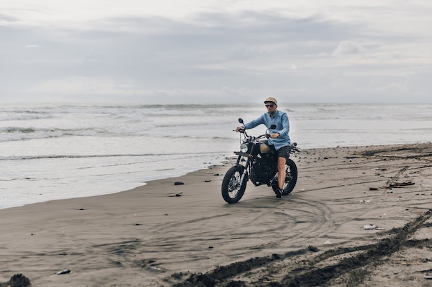 Mężczyzna w czapce, jazda motocyklem na plaży. Moto cross dirtbiker na plaży o zachodzie słońca na Bali. Młody hipster mężczyzna cieszący się wolnością i aktywnym trybem życia, bawiąc się na wycieczce motocyklowej.