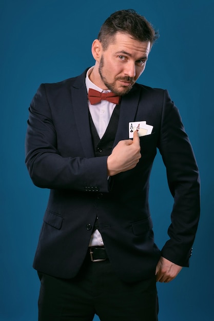 Mężczyzna w czarnym klasycznym garniturze i czerwonej muszce, pokazujący dwie karty do gry, pozujący na tle niebieskiego studia...