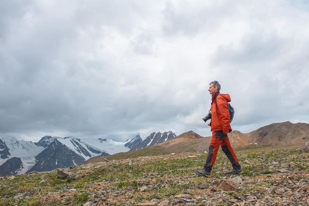 Mężczyzna w czerwieni z aparatem spaceruje z widokiem na duże ośnieżone pasmo górskie z ostrymi wierzchołkami pod szarym pochmurnym niebem Dramatyczny krajobraz z turystą w wysokich górach Śnieżne spiczaste szczyty przy zmiennej pogodzie