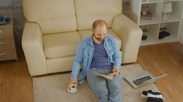 Mężczyzna W Dżinsowej Koszuli śmiejący Się Podczas Czytania Książki I Picia Kawy Na Podłodze