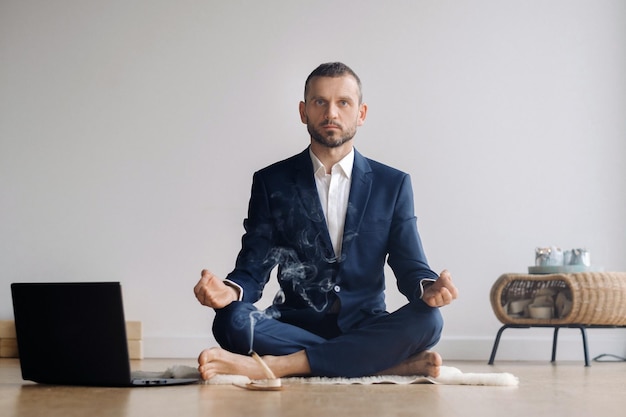 Mężczyzna w formalnym garniturze medytuje siedząc w sali fitness z laptopem