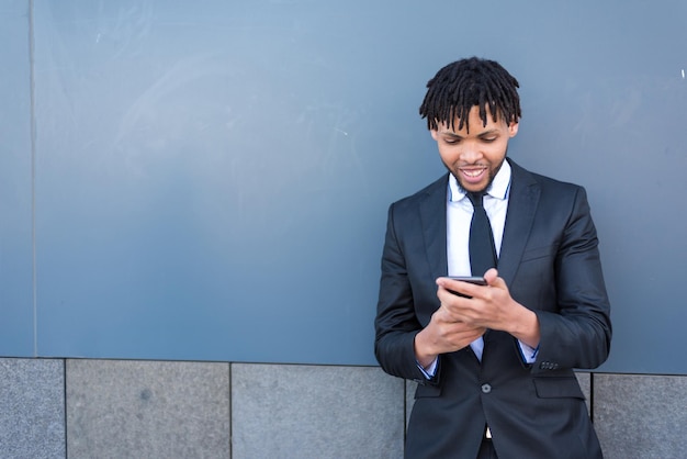 Mężczyzna w garniturze na ulicy wysyła SMS-y na smartfonie i uśmiecha się, odwracając wzrok w niebieskiej ścianie obszaru biznesowego