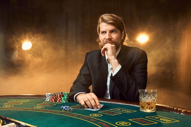 Mężczyzna w garniturze siedzący przy stole do gry. Mężczyzna gracz. Pasja, karty, żetony, alkohol, kości, hazard, kasyno - to jak męska rozrywka.
