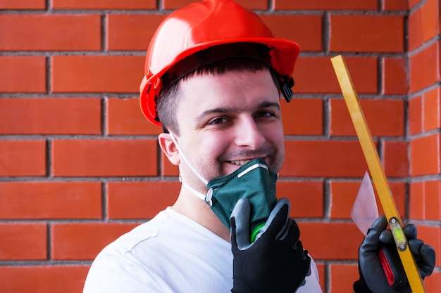 Mężczyzna w hełmie ochronnym nosi respirator na tle czerwonej ściany z cegły.