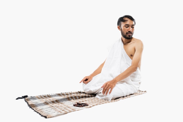 Mężczyzna w ihramie modlący się z dywanem modlitewnym podczas wykonywania salam na odosobnionym tle