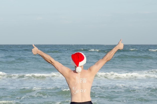 Mężczyzna w kapeluszach Mikołaja z napisem Nowy Rok z tyłu na plaży Kciuki w górę Widok z tyłu