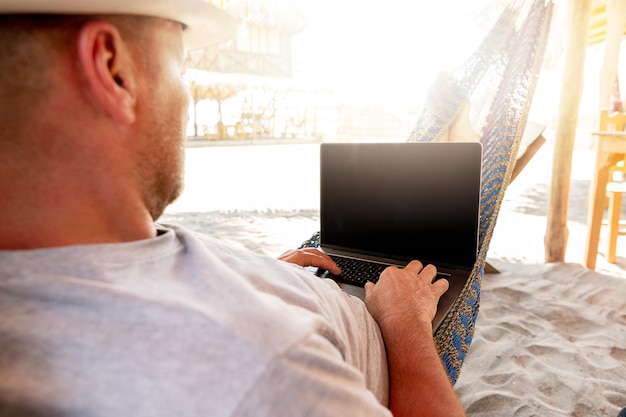 Mężczyzna w kapeluszu zdalnie korzysta z laptopa na plaży w pobliżu domku Playa La Ventanilla