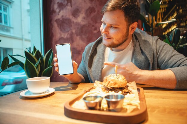 Mężczyzna w kawiarni trzymający telefon z białym ekranem je burger pijący herbatę