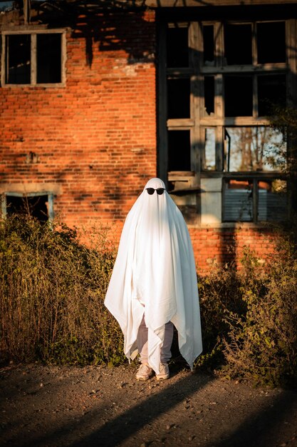 Zdjęcie mężczyzna w kostiumie ducha wykonanym z prześcieradła i okularów przeciwsłonecznych stoi w pobliżu opuszczonego budynku ghost challenge 2021 spooky season celebrating halloween