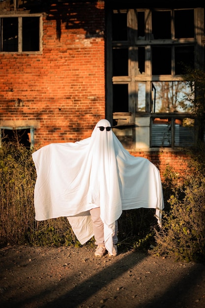 Mężczyzna w kostiumie ducha wykonanym z prześcieradła i okularów przeciwsłonecznych stoi w pobliżu opuszczonego budynku Ghost Challenge 2021 Spooky season Celebrating halloween