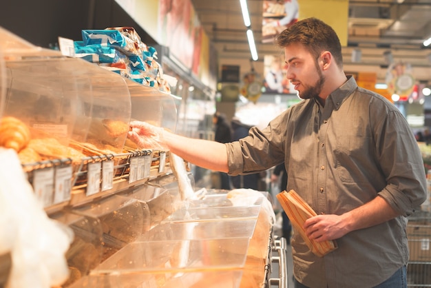 Mężczyzna w koszuli trzyma w rękach papierową torbę, stoi w sklepie z chlebem w supermarkecie i wybiera bułki.