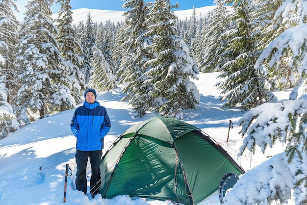 Mężczyzna w lesie w zimowych górach z głębokim śniegiem, stojący w pobliżu namiotu i patrzący w kamerę