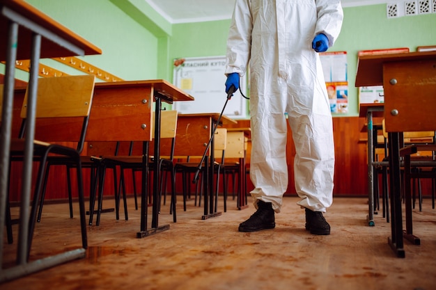 Mężczyzna w ochronnym kombinezonie antybakteryjnym sprząta klasę sprayem z płynem odkażającym. Profesjonalny pracownik sanitarny dezynfekuje audytorium specjalnymi urządzeniami. Pojęcie opieki zdrowotnej.