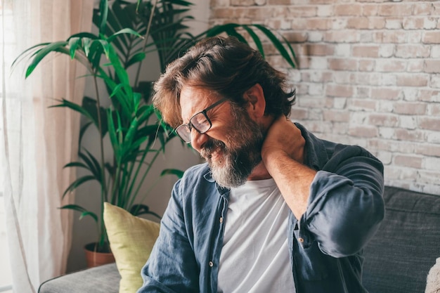 Mężczyzna w okularach trzymający się za bolącą szyję siedzący na kanapie w domu Kaukaski facet cierpiący na ból szyi z powodu nieprawidłowej postawy lub zapalenia stawów Sfrustrowany dojrzały mężczyzna z silnym bólem