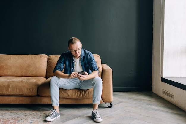 Zdjęcie mężczyzna w okularach w zwykłych ubraniach, siedzący na kanapie, surfujący po internecie na smartfonie