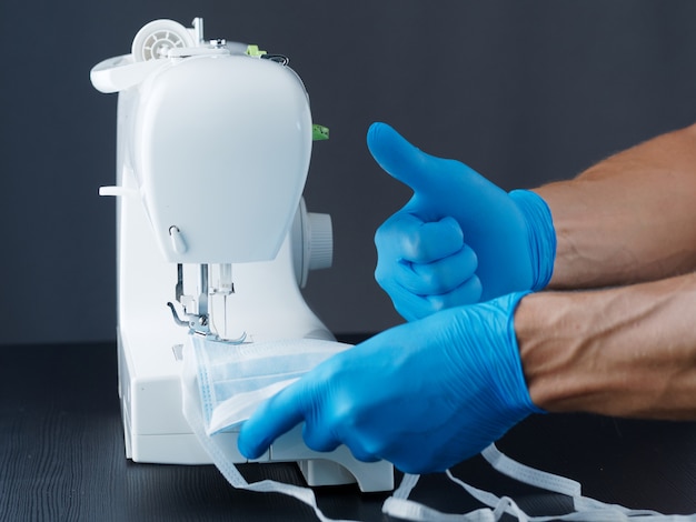 Mężczyzna w rękawiczkach robi maski medyczne na maszynie do szycia i pokazuje kciuki do góry.