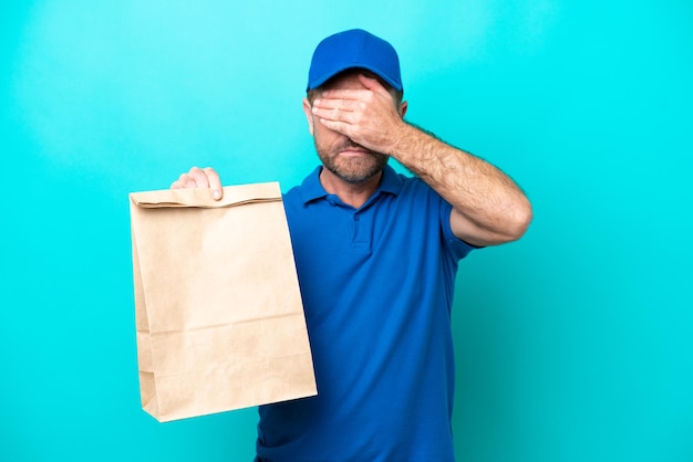 Mężczyzna w średnim wieku biorący torbę jedzenia na wynos odizolowany na niebieskim tle zakrywający oczy rękami Nie chce czegoś widzieć