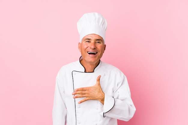 Mężczyzna w średnim wieku kucharz na białym tle śmieje się głośno, trzymając rękę na piersi.