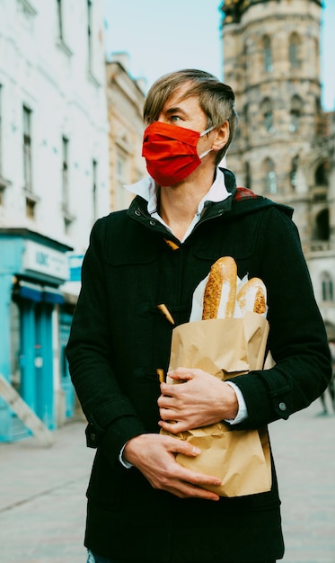 Zdjęcie mężczyzna w średnim wieku na ulicy z chlebem, bagietką, zakupami bochenków podczas globalnej pandemii, w masce, kupując chleb z piekarni. jedzenie na wynos, zakupy w piekarni podczas covid 19.