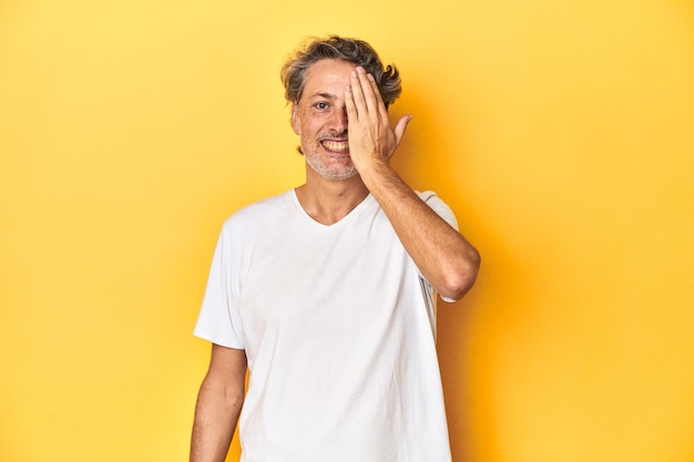 Mężczyzna w średnim wieku pozuje na żółtym tle, bawiąc się, pokrywając połowę twarzy dłonią