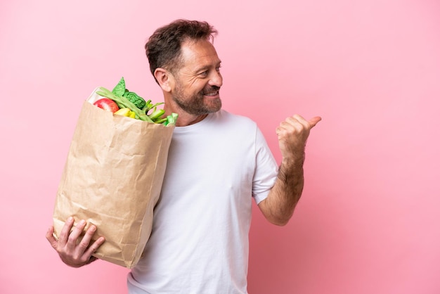 Mężczyzna w średnim wieku trzymający torbę na zakupy spożywcze na białym tle na różowym tle wskazujący na bok, aby zaprezentować produkt