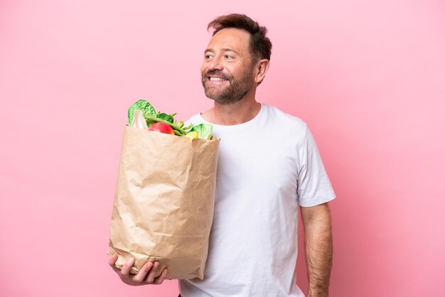 Mężczyzna w średnim wieku trzymający torbę na zakupy spożywcze odizolowaną na różowym tle, myślący o pomyśle, patrząc w górę