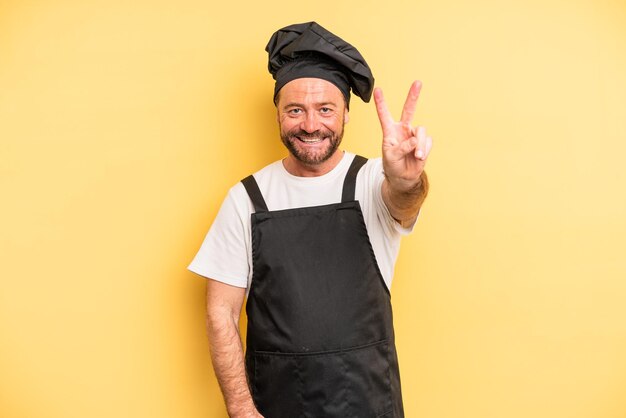 Mężczyzna w średnim wieku uśmiechający się i wyglądający na szczęśliwego, gestykulując zwycięstwo lub koncepcję szefa kuchni pokoju