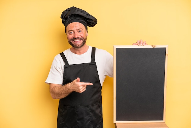 Mężczyzna w średnim wieku uśmiechający się radośnie, czując się szczęśliwy i wskazując na bok. koncepcja szefa kuchni i tablicy