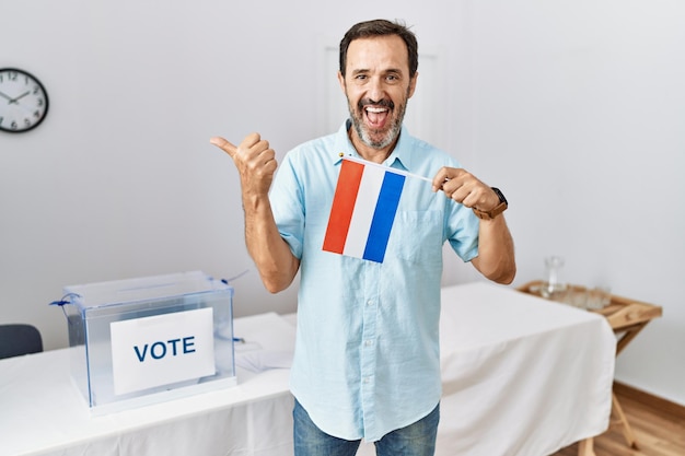 Mężczyzna w średnim wieku z brodą podczas wyborów w kampanii politycznej trzymający flagę holandii wskazujący kciuk na bok uśmiechnięty zadowolony z otwartymi ustami