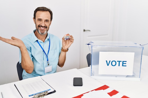 Zdjęcie mężczyzna w średnim wieku z brodą siedzi przy karcie do głosowania, trzymając odznakę głosuję, uśmiechając się wesoło, przedstawiając i wskazując dłonią patrząc w kamerę