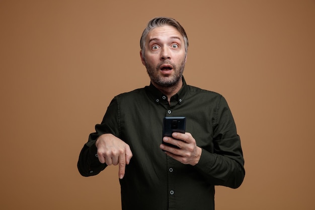 Mężczyzna w średnim wieku z siwymi włosami w ciemnej koszuli trzymający smartfona patrzący w kamerę zdumiony i zaskoczony, wskazując palcem wskazującym w dół, stojący na brązowym tle