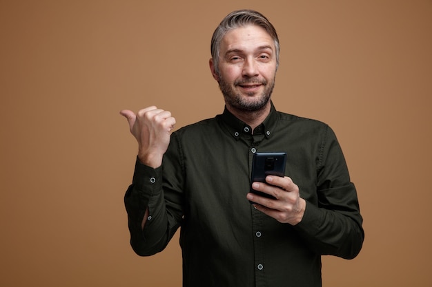 Mężczyzna w średnim wieku z siwymi włosami w ciemnej koszuli, trzymający smartfona wskazującego kciukiem w bok, uśmiechający się pewnie, stojący na brązowym tle