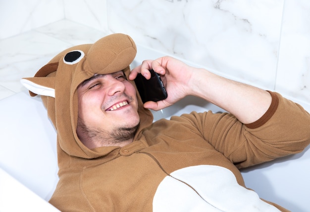 Mężczyzna w stroju cosplay krowy. Facet w zabawnej zwierzęcej piżamie leży w wannie i rozmawia przez telefon komórkowy.