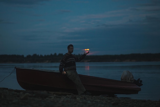 Mężczyzna w swetrze robi selfie na tle łodzi nad rzeką w mistyczną księżycową noc