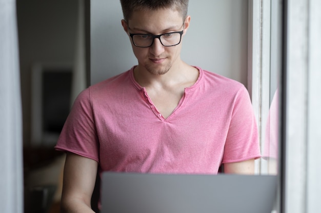 mężczyzna w szortach i różowej koszulce siedzi na oknie i pracuje na laptopie