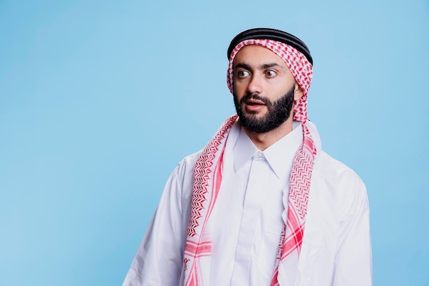 Mężczyzna w tradycyjnych ubraniach muzułmańskich odwrócił wzrok zszokowany wyraz twarzy Arab ubrany w thobe i chustę stojący z zaskoczonymi emocjami i otwartymi ustami podczas pozowania w studio