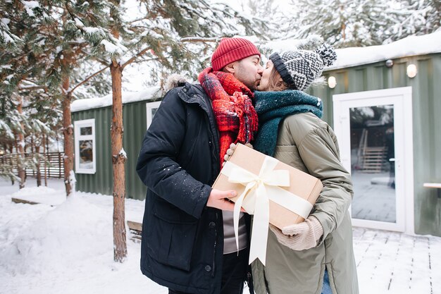 Mężczyzna w zimowych ubraniach całuje swoją kobietę, po podarowaniu jej prezentu. Stojąc przed małym zaśnieżonym domem.